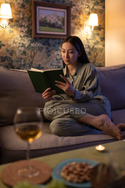 Barefoot Asian female resting on sofa and enjoying reading in evening at home — Stock Photo