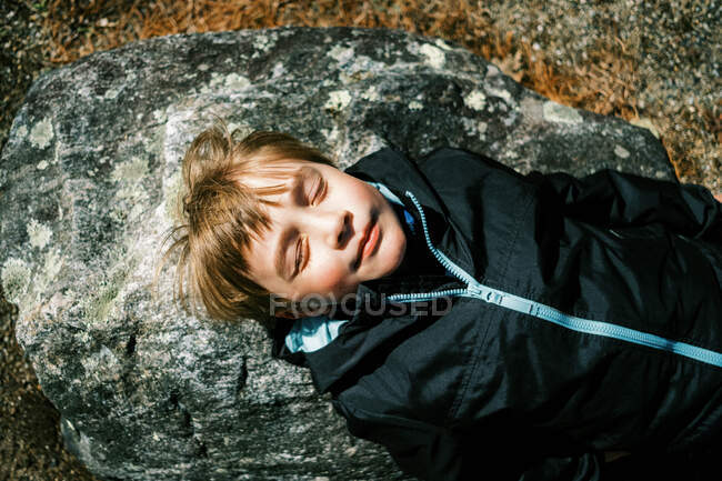 Niño acostado en la roca y disfrutando de los primeros rayos de sol cálidos en la cara - foto de stock