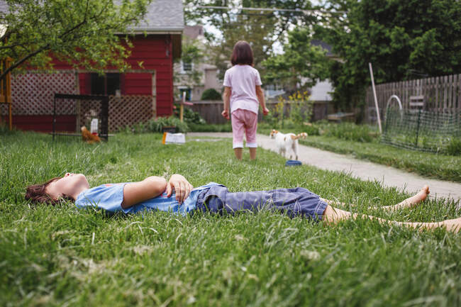 Un niño se acuesta en hierba verde en el patio con la hermana y el gato en el fondo - foto de stock