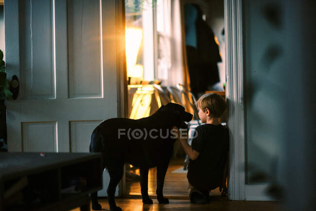 Маленький мальчик ласкает свою собаку у окна с закатом на заднем плане — стоковое фото