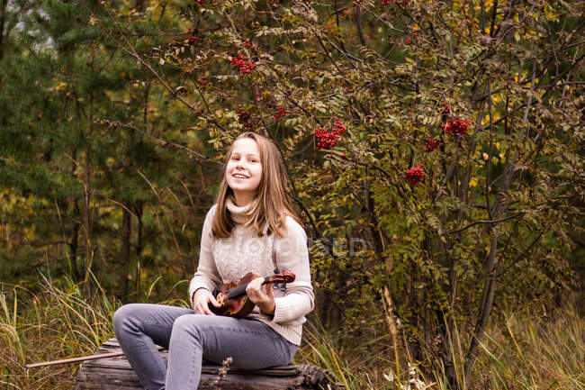 Ein schönes Teenager-Mädchen mit langen blonden Haaren sitzt auf einem Baumstamm im Park und hält eine Geige in den Händen, sieht happ aus — Stockfoto