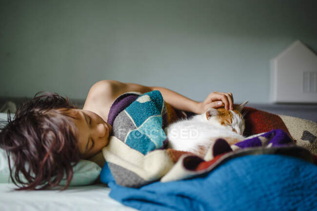 Uma criança deita na cama sob uma pilha de cobertores aconchegantes aconchegantes com um gato — Fotografia de Stock
