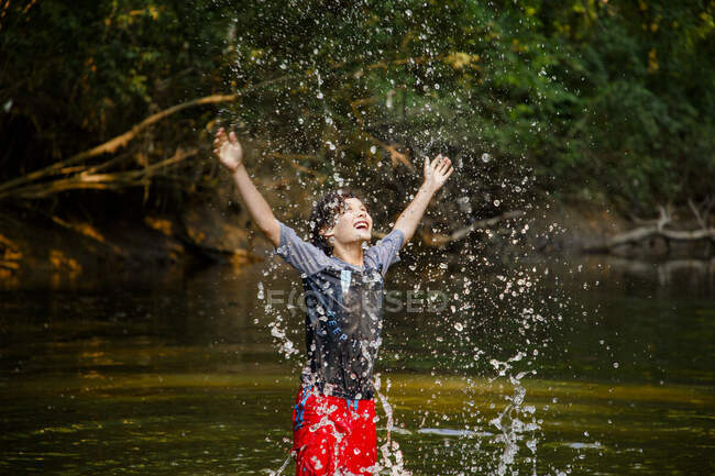 A joyful boy playing in river flings water drops high into the air — Stock Photo