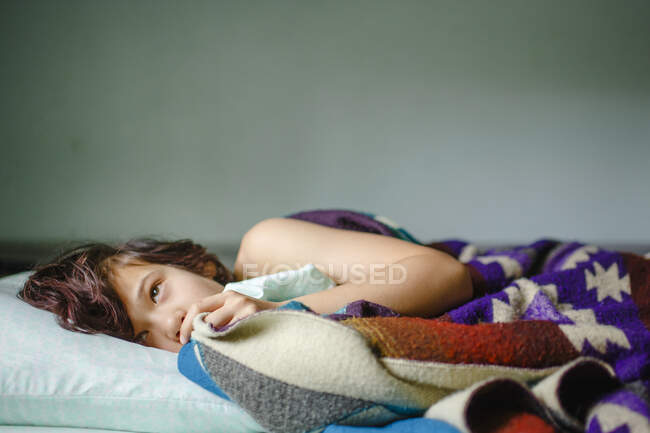 Um menino pequeno deita-se na cama olhando para cima envolto em cobertor colorido — Fotografia de Stock