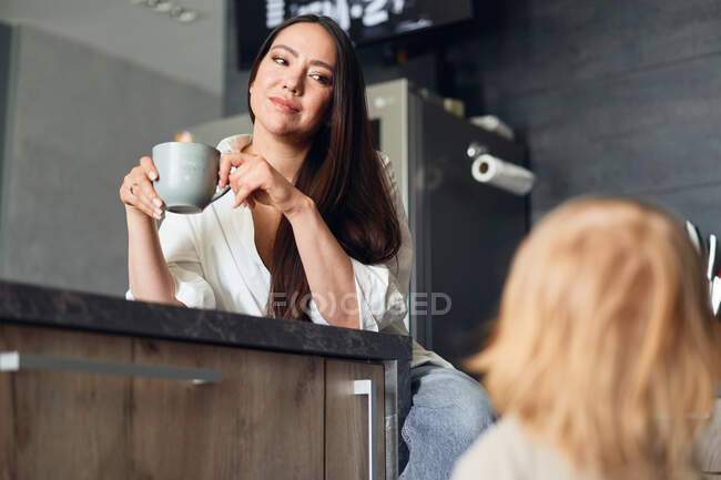 Mulher jovem sentada na cozinha com xícara de café e olhando para o filho — Fotografia de Stock