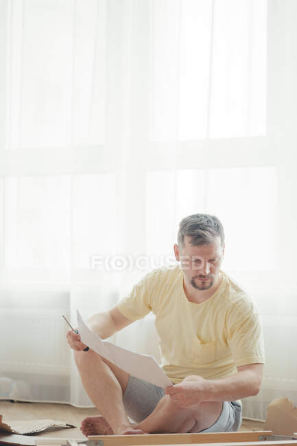Молода приваблива людина в жовтій футболці складає меблі відповідно до інструкцій, сидячи в світлій і повітряній вітальні. Збірка меблів вдома. Самоізоляція, саморобна . — стокове фото