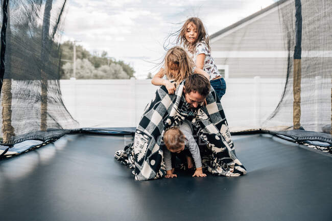 Três crianças escalando o pai enquanto jogam em um trampolim em casa — Fotografia de Stock