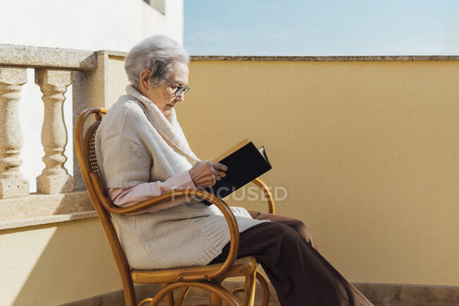 Donna anziana che legge su una terrazza soleggiata all'aperto — Foto stock