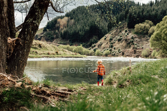Pequena criança encaracolada explorando margem do rio em botas — Fotografia de Stock