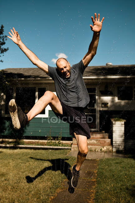 Un hombre de mediana edad saltando a través de un aspersor en el verano - foto de stock