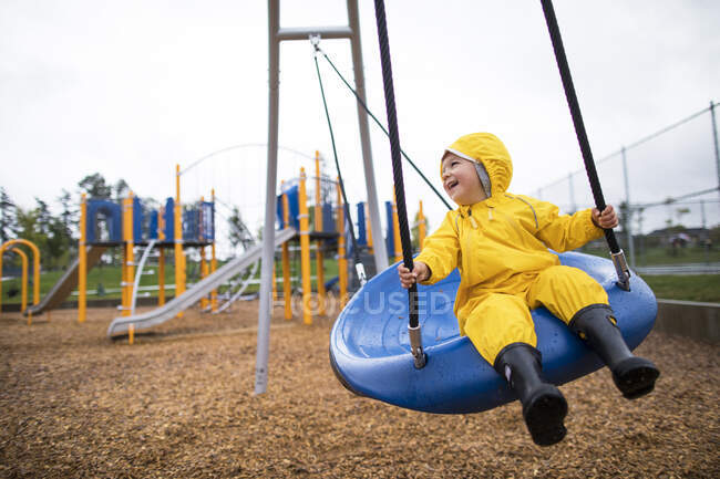 Menino feliz monta no balanço no parque em um dia molhado — Fotografia de Stock