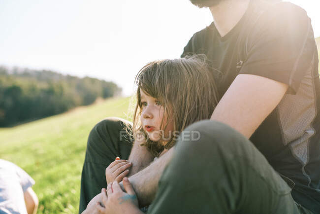 Niña apoyada contra su padre mientras descansa en una colina bajo el sol - foto de stock