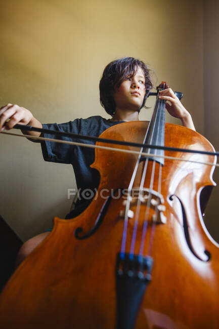 Мальчик с серьезным выражением лица играет на виолончели, глядя в окно — стоковое фото
