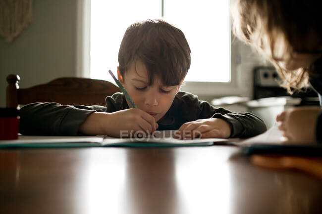 Мальчик делает домашнее задание карандашом за кухонным столом с сестрой — стоковое фото