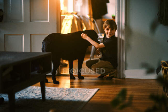 Petit garçon caressant son chien par une fenêtre avec coucher de soleil en arrière-plan — Photo de stock
