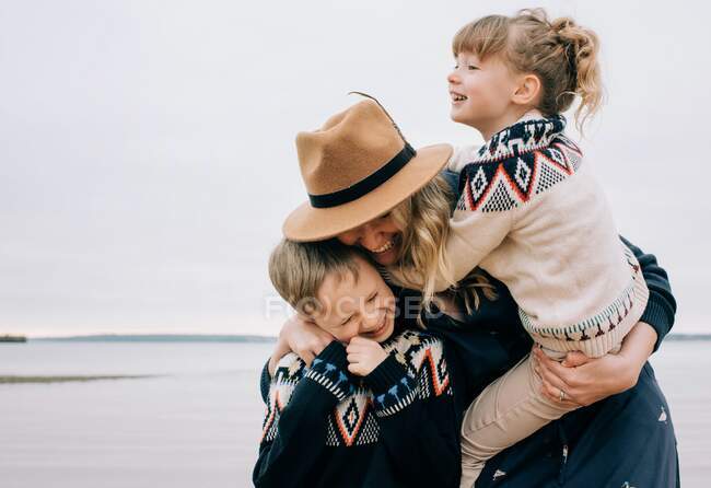 Mom hugging her kids laughing whilst walking along the beach — Stock Photo