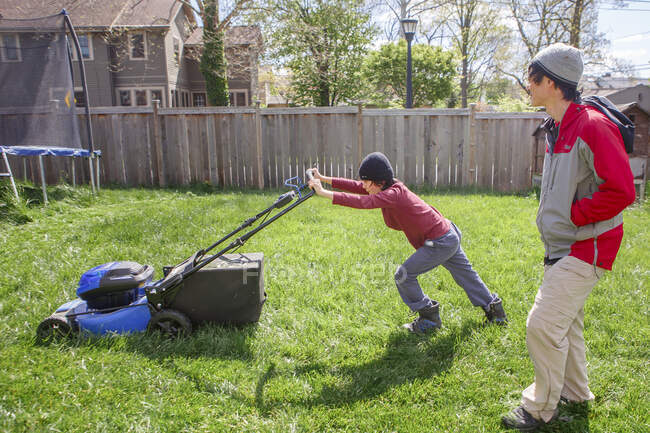 Menino trabalha duro para empurrar cortador de grama elétrico, enquanto o pai assiste em — Fotografia de Stock