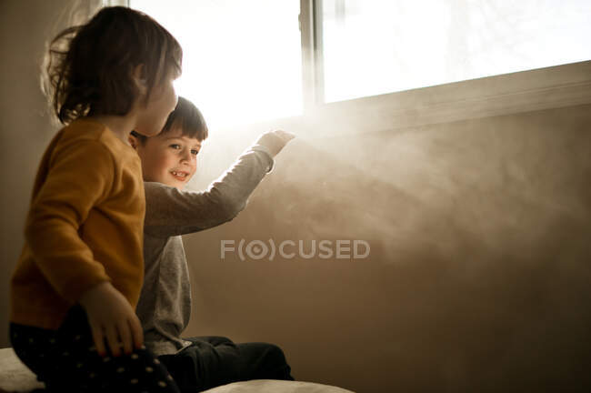 A brother and sister sitting on a bed watching rays of light — Stock Photo