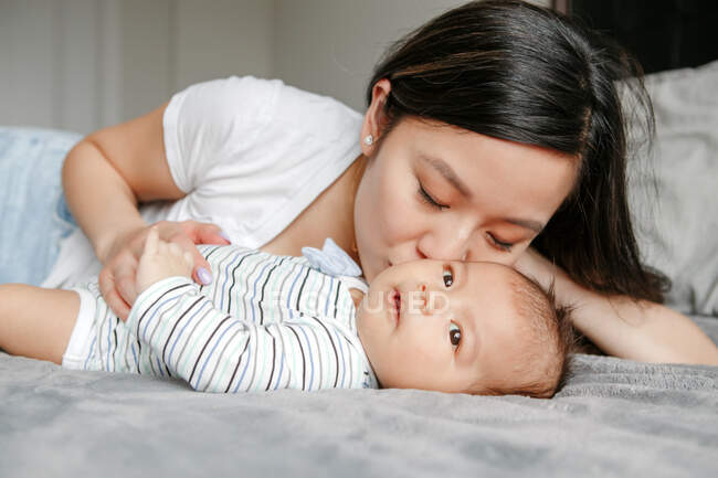 Vacaciones del día de las madres. Orgullosa sonriente madre asiática china abrazando a un bebé recién nacido besándose hijo. Familia feliz acostada en la cama en el dormitorio. Hogar estilo de vida auténtico momento. Diversidad étnica. - foto de stock