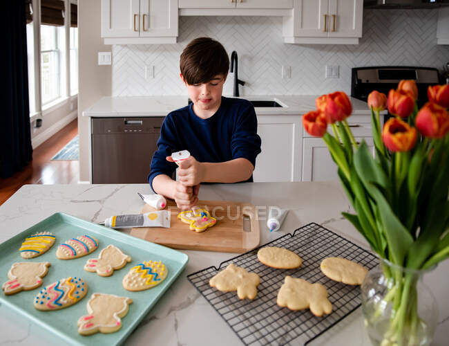 Menino jovem que decora biscoitos de Páscoa no balcão de uma cozinha moderna. — Fotografia de Stock