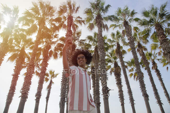 Latino na casa dos 20 anos a tirar uma selfie entre as palmeiras da praia. Homem de pele escura tirando uma selfie de si mesmo enquanto está de pé. — Fotografia de Stock