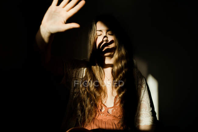 Una mujer levantando su mano a la luz proyectando una sombra sobre su rostro - foto de stock
