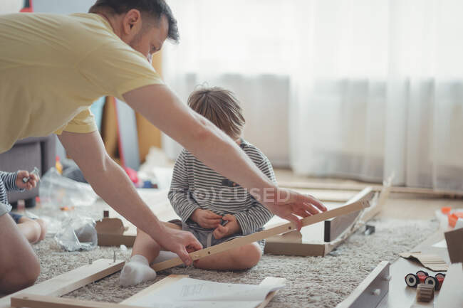 Padre e figlio stanno assemblando mobili un tavolo di legno secondo le istruzioni in un luminoso soggiorno. Interni domestici e domestici. Una famiglia. Mani di un uomo e di un bambino con uno strumento. Primo piano. — Foto stock