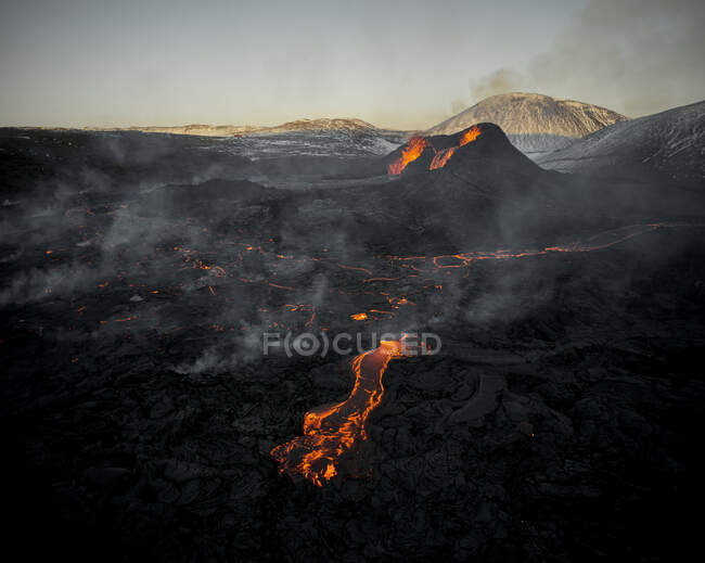 Espectacular paisaje de montaña volcánica en erupción con magma naranja caliente que fluye en la superficie rocosa al atardecer - foto de stock