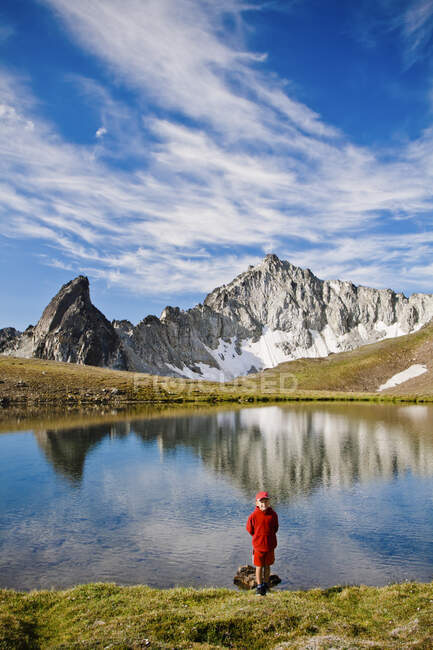 Young boy wearing red, standing in front of alpine lake, scenic peaks — Stock Photo