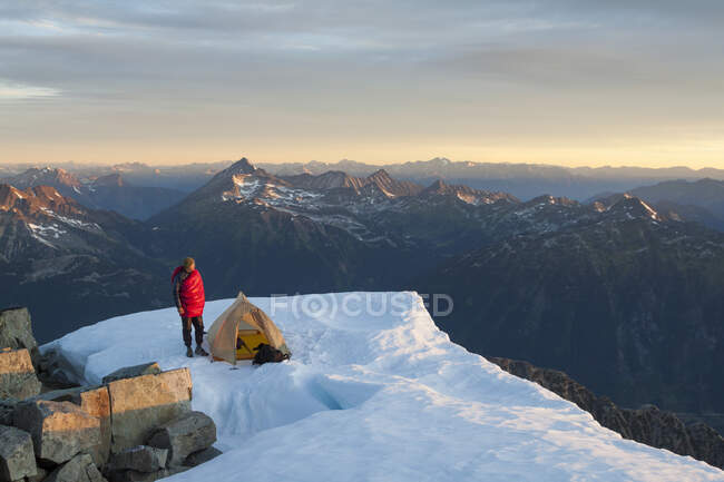 Un escalador está de pie junto a su tienda, encaramado en la cima de una montaña. - foto de stock