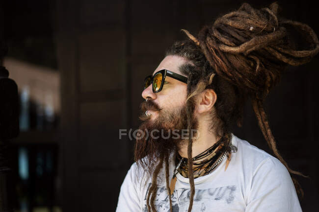 Retrato de un tipo hipster con gafas y con rastas y un tatuaje se sienta en el fondo de una casa de madera - foto de stock