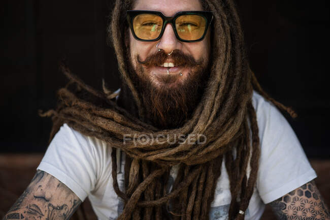 Retrato de um cara hipster com óculos e com dreadlocks e uma tatuagem se senta no fundo de uma casa de madeira — Fotografia de Stock