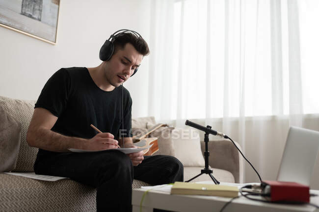 Человек в наушниках сидит на диване и пишет заметки, создавая музыку дома — стоковое фото