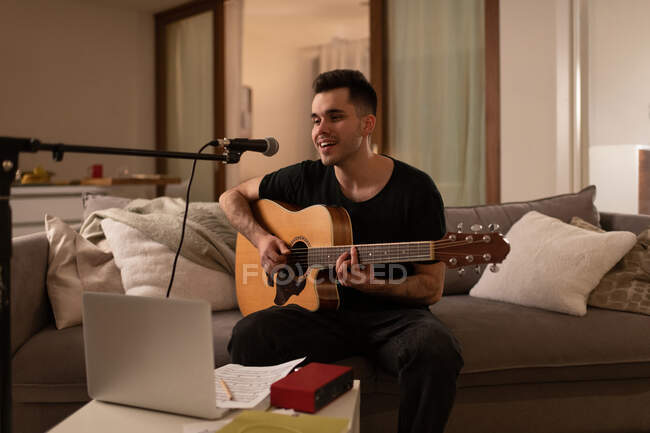 Cara feliz tocando guitarra acústica e cantando enquanto sentado no sofá em casa — Fotografia de Stock