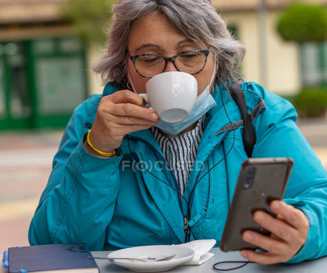 Femme aux cheveux blancs boire du café au bar tout en écrivant et en regardant le téléphone — Photo de stock