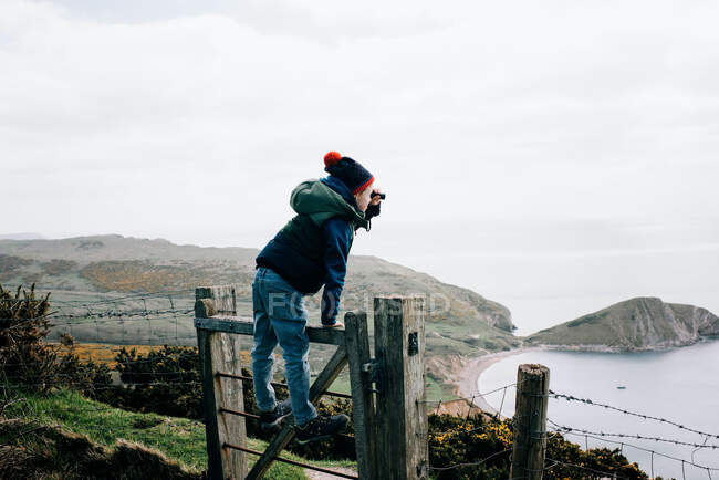 Boy stood on a fence looking through binoculars at the Jurassic coast — Stock Photo