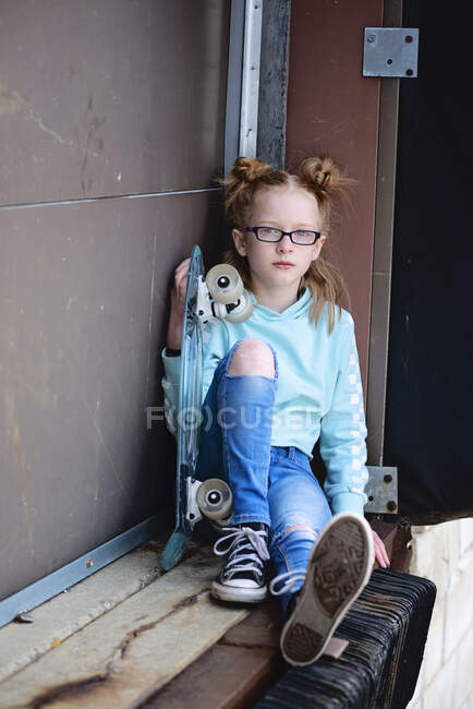 Tween menina com cabelo vermelho e skate sentado em uma doca de carregamento. — Fotografia de Stock