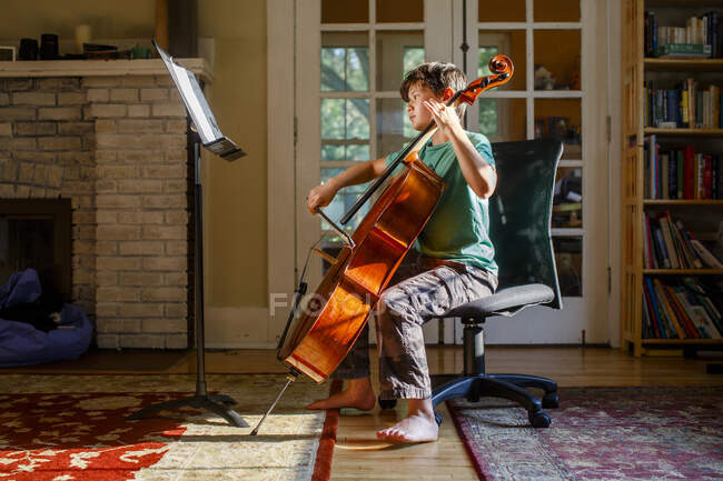 Un chico descalzo practica violonchelo con luz de ventana dorada en interiores - foto de stock