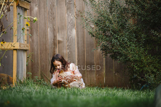 Una linda niña juega con un pollo en un patio lleno de flores - foto de stock