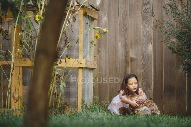 A beautiful little girl kneels down in grass to pick up a chicken — Stock Photo