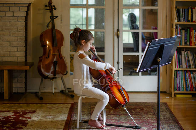 Uma criança graciosa focada pratica violoncelo na luz da janela em casa — Fotografia de Stock