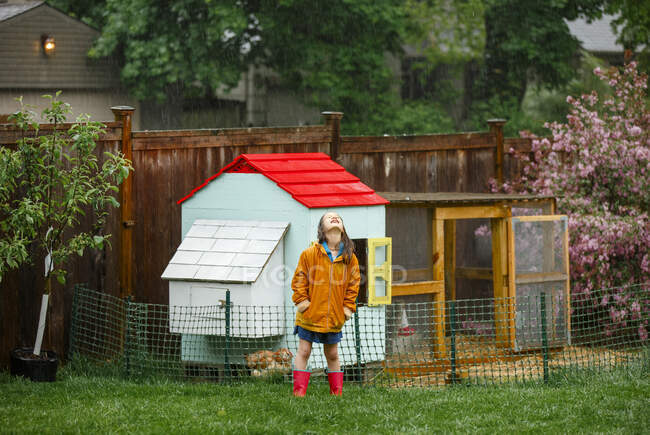 A joyful child stands in the rain in garden by chicken coop looking up — Stock Photo
