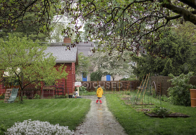 Rear-view of child standing in rain on path through backyard garden — Stock Photo