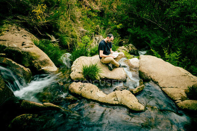 Hombre leyendo un libro sentado en una roca junto a un río - foto de stock