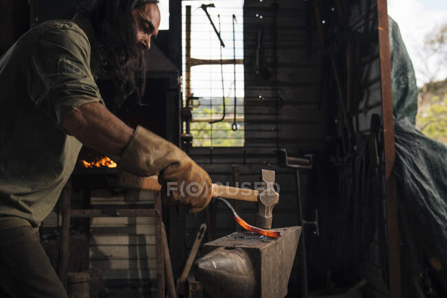 Herrero trabajando un pedazo de acero al rojo vivo con un martillo. - foto de stock