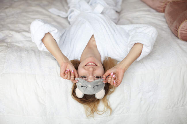 Young attractive smiling woman in sleep mask and bathrobe lie on bed, looking at camera — Stock Photo
