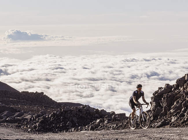 Male bicyclist rides up gravel road above clouds on Mauna Kea, Hawaii — Stock Photo