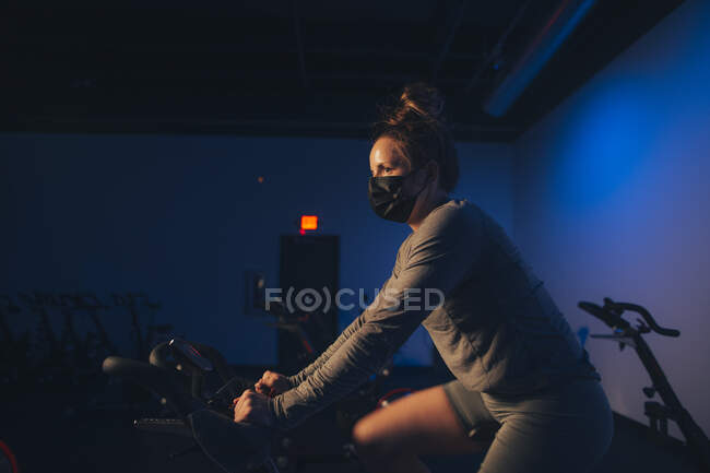 Uma jovem mulher branca pedala em um estúdio interno com uma máscara. — Fotografia de Stock