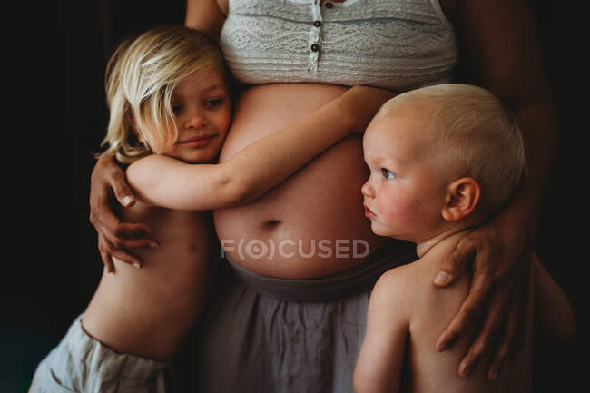 Crianças loiras bonitas abraçando a barriga grande grávida da mãe em casa — Fotografia de Stock