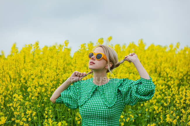 Belle blonde aux lunettes de soleil jaunes et robe verte sur champ de colza jaune — Photo de stock